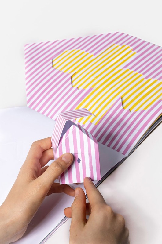 hands-fold-and-assemble-pink-paper-cabin